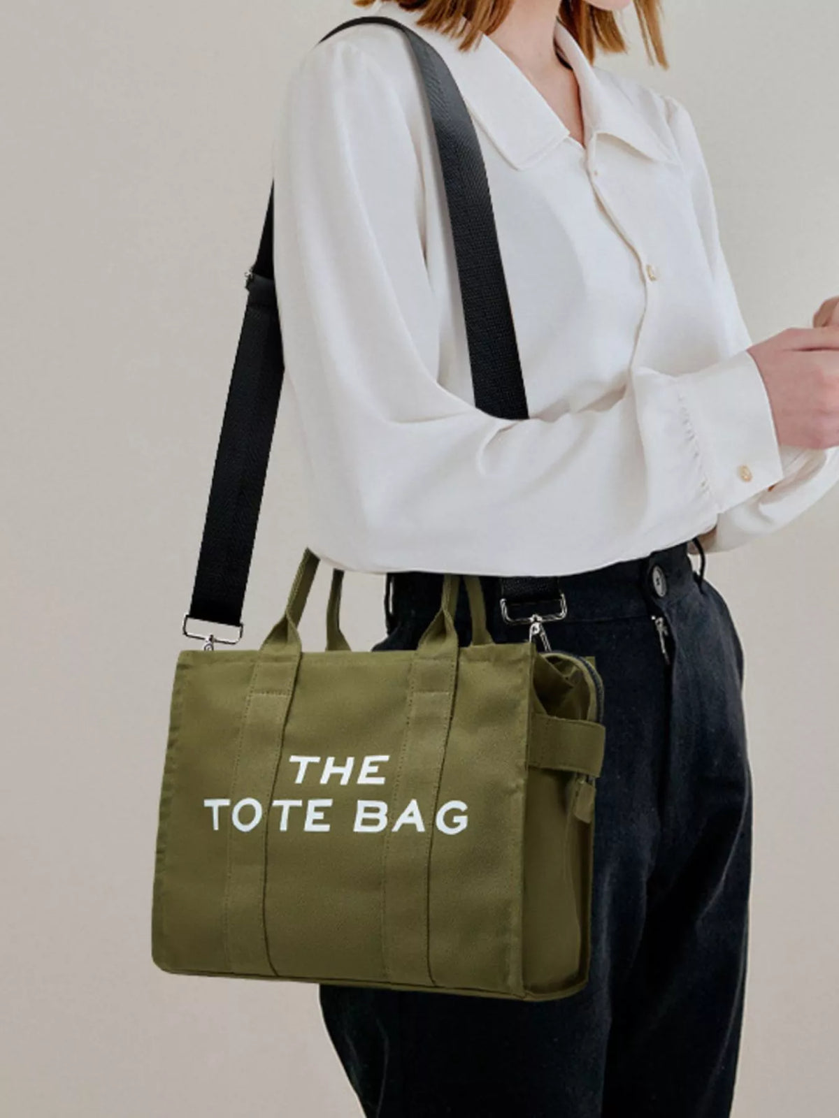 Army green canvas tote bag with “THE TOTE BAG” lettering, worn with long shoulder strap