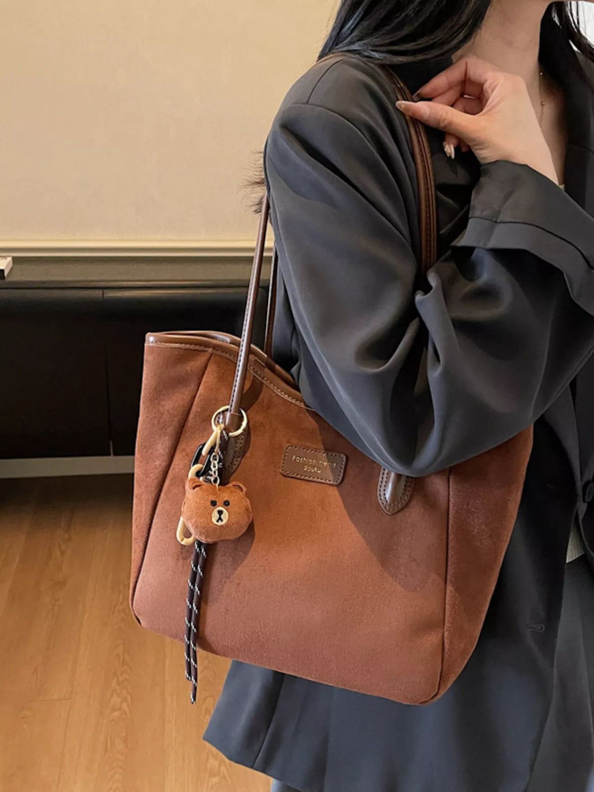Brown suede tote bag with long handles, front logo patch, and bear keychain tassel
