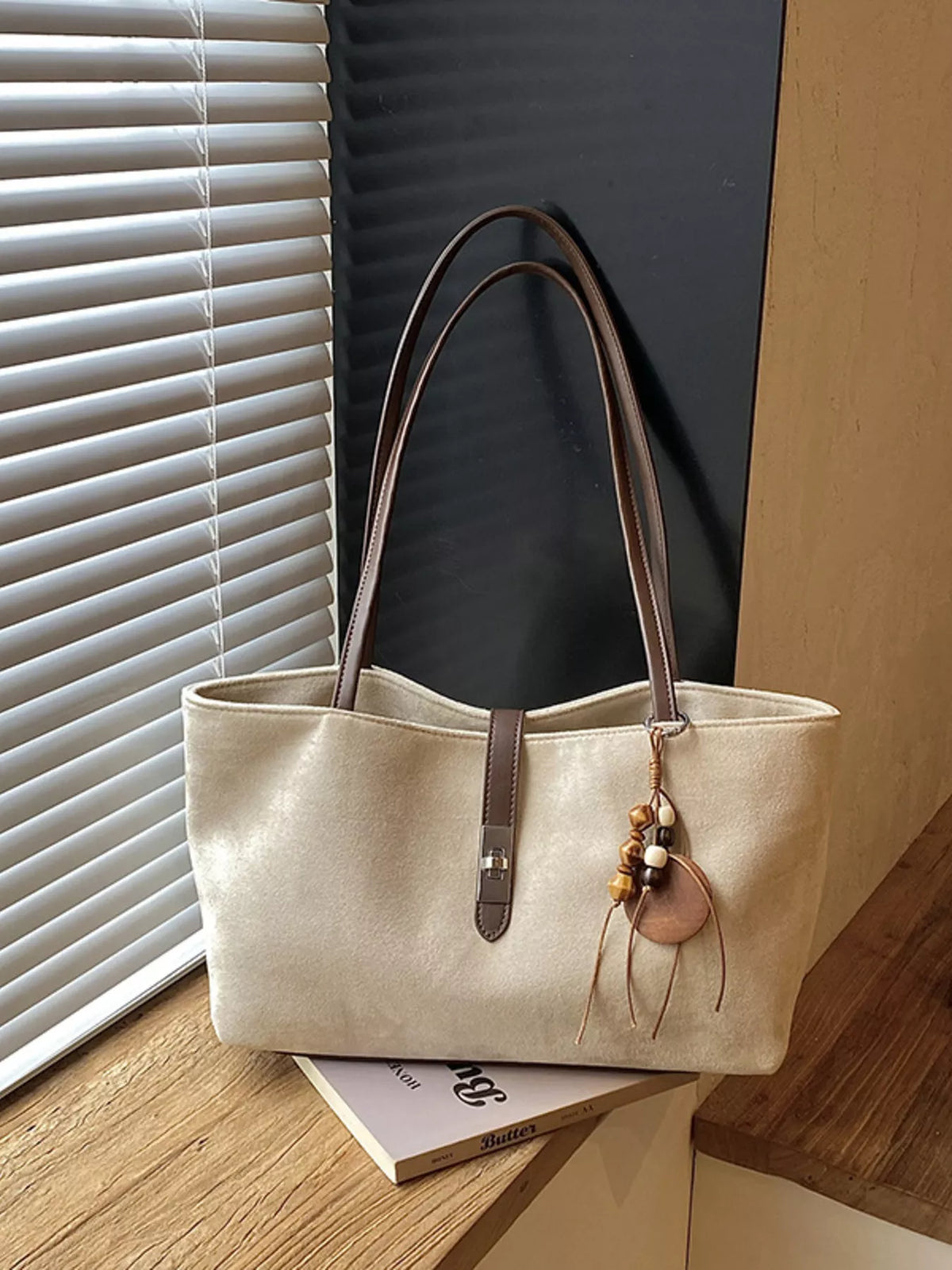 Beige suede tote bag with top handles, vertical buckle strap, and hanging charm