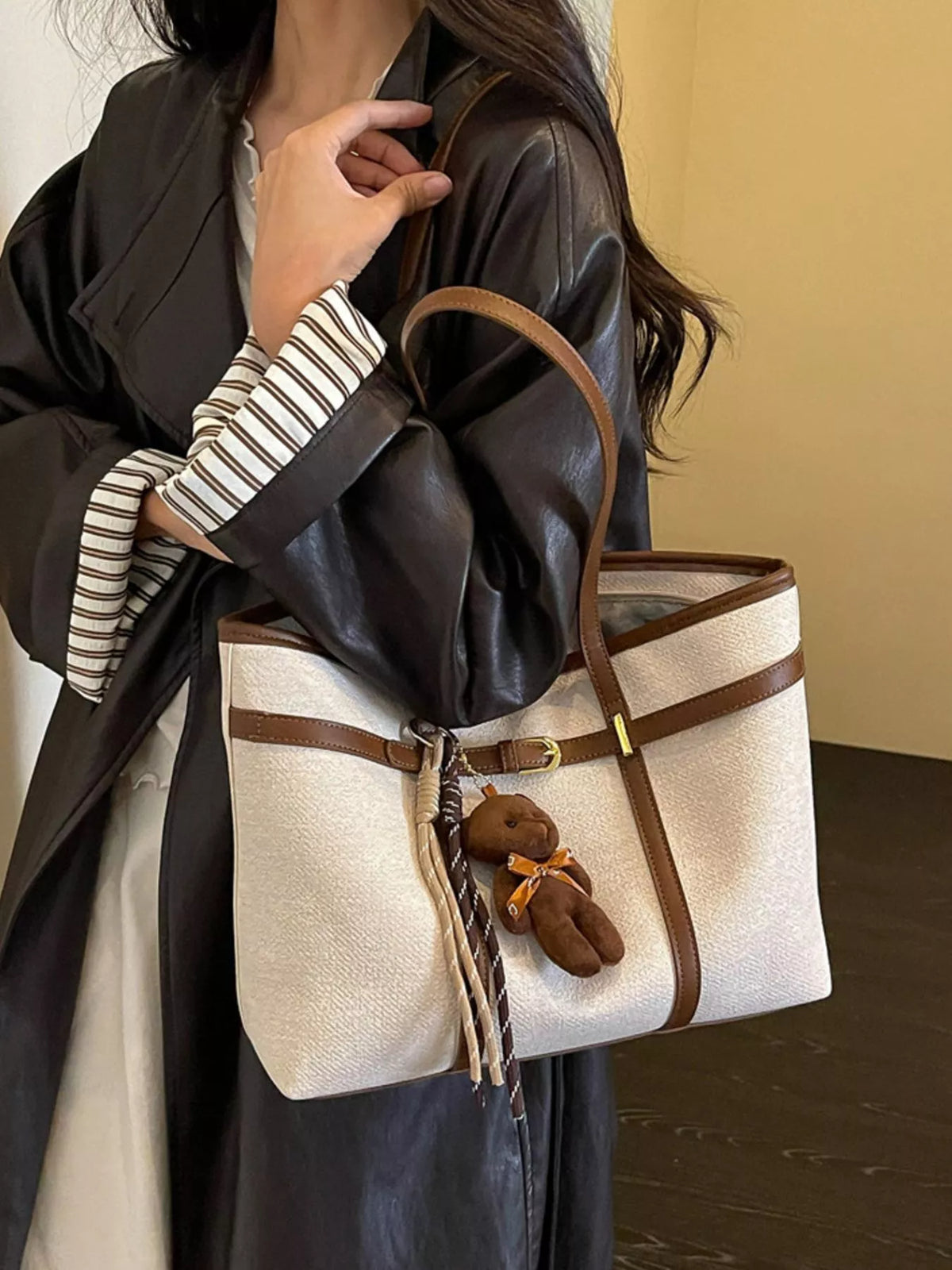 Off-white tote bag with brown top handles, buckle center strap, and teddy charm