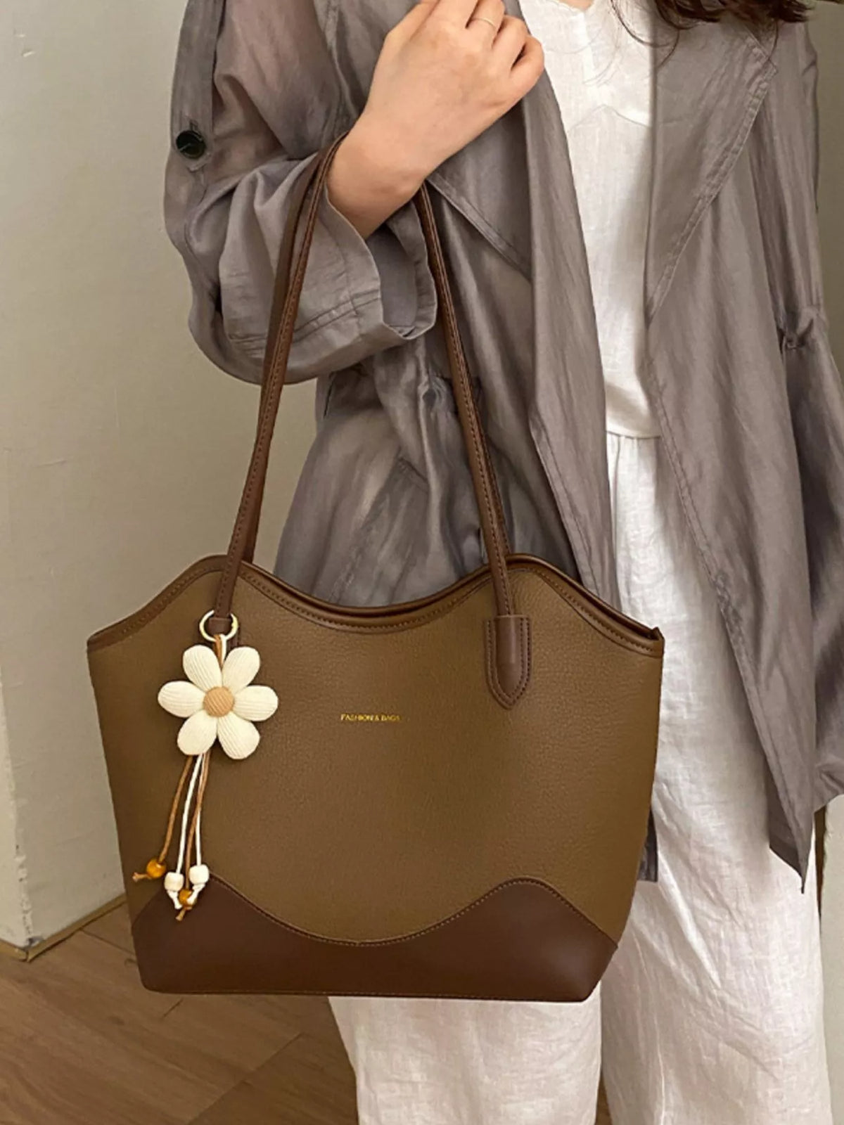 Brown vegan leather shoulder bag with top handles, curved front seam, and flower tassel charm