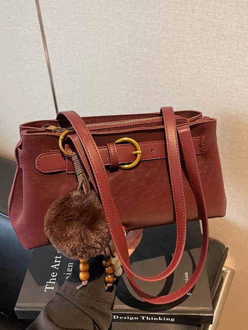Burgundy buckle-detail shoulder bag with double top handles, zipper top, and pom-pom