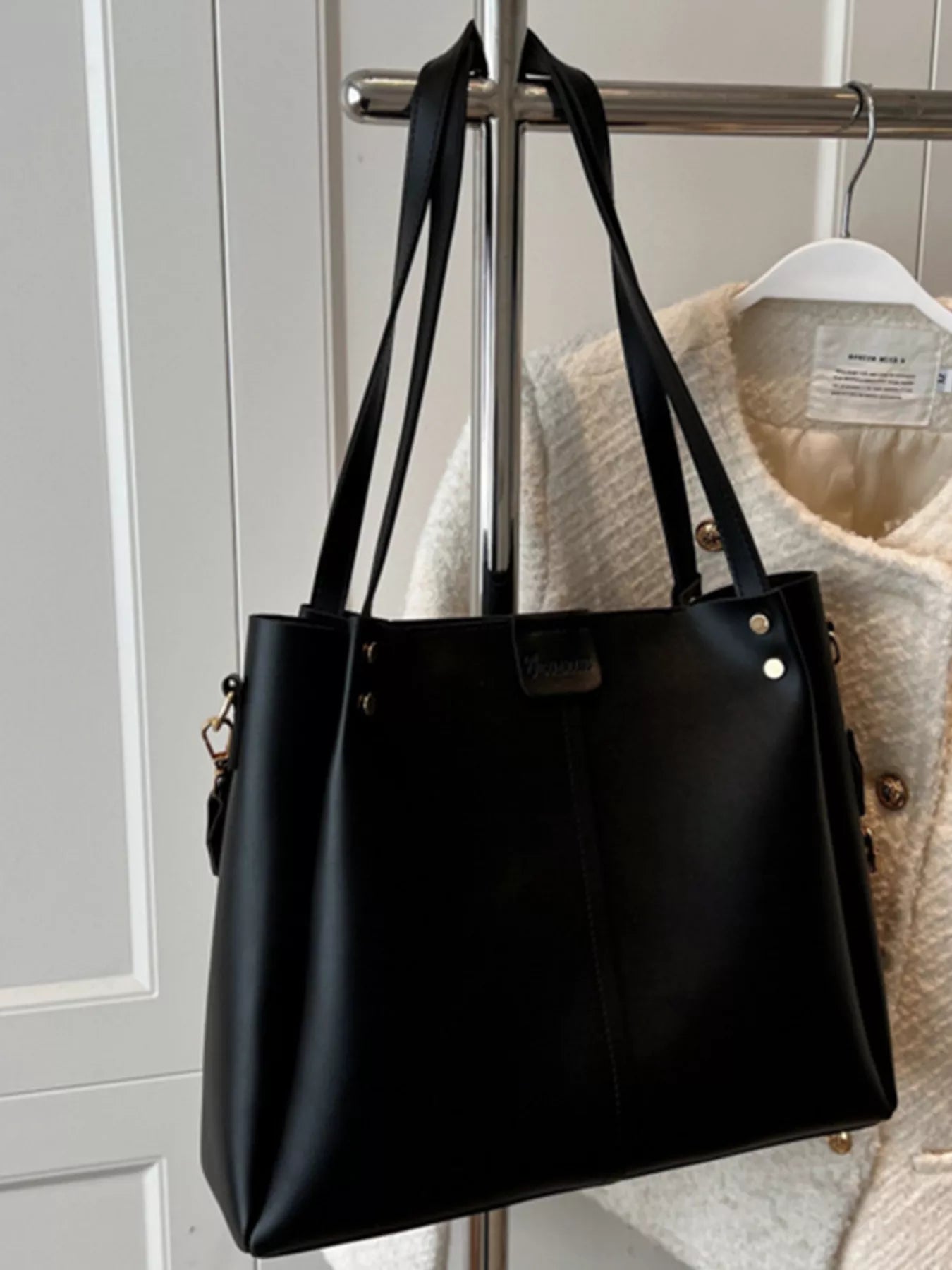 Black tote-style shoulder bag with wide straps, metal feet, and small front patch