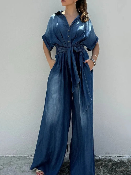 Blue denim jumpsuit with button front, tie waist belt, and wide-leg trousers.