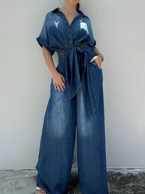 Blue denim button-front jumpsuit with waist tie belt and wide-leg pant legs.