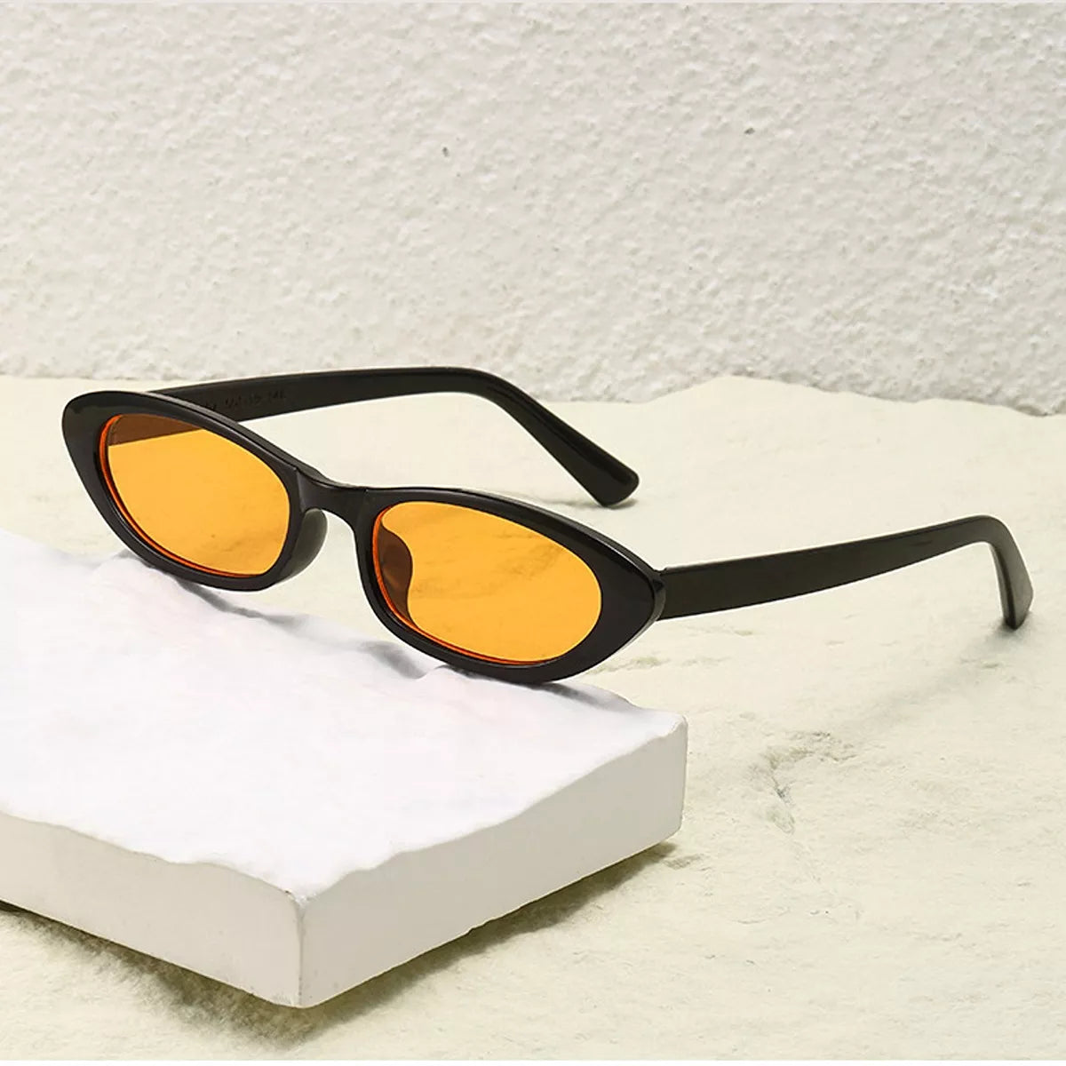 Black oval UV400 sunglasses with amber lenses and curved arms on a white block