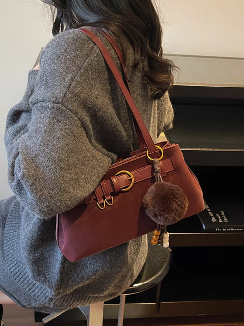 Burgundy buckle-detail shoulder bag with front belt strap, gold rings, and pom-pom charm
