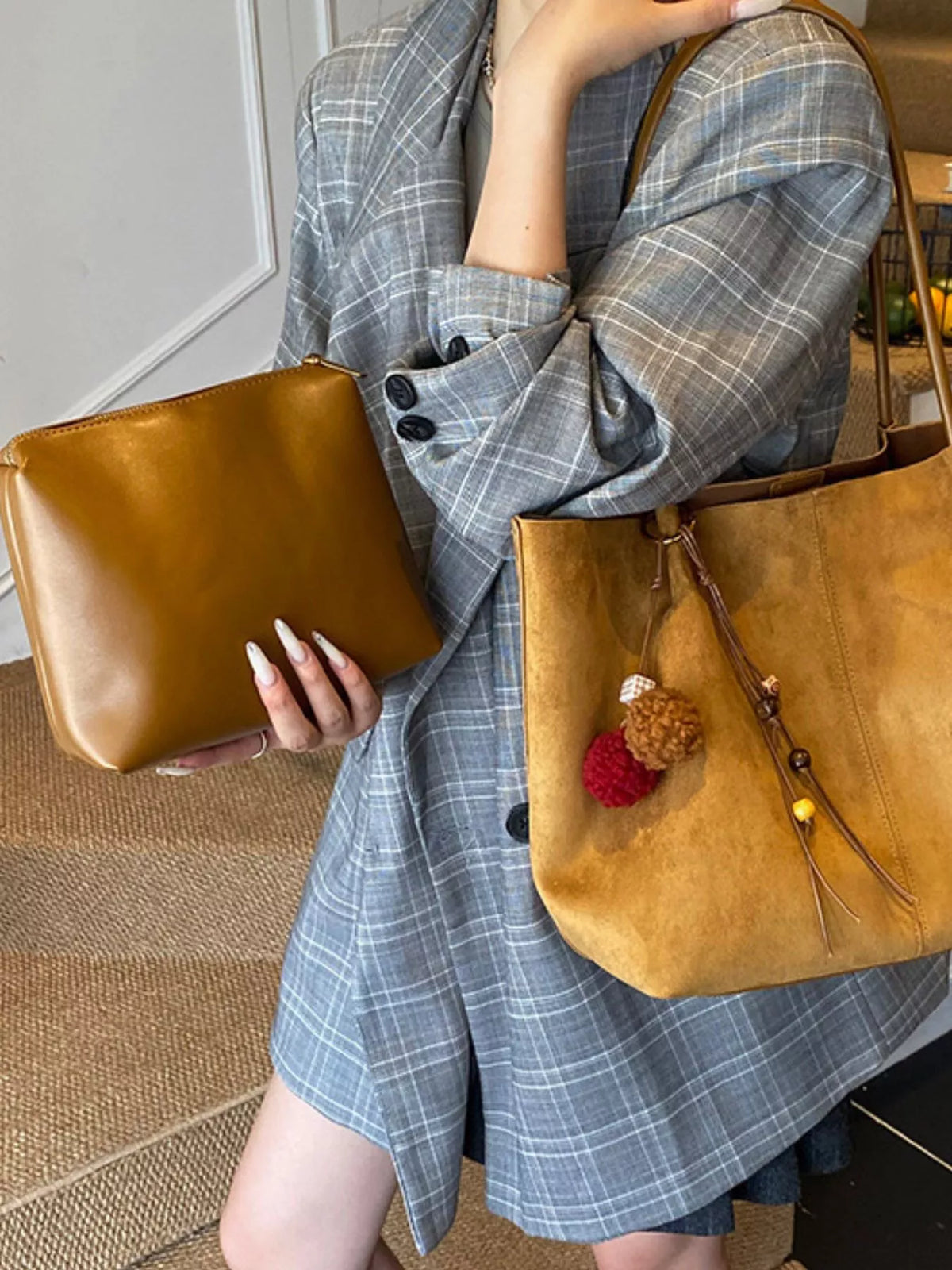 Tan suede tote bag held by model; long handles, front zip pocket, and pom-pom charm.