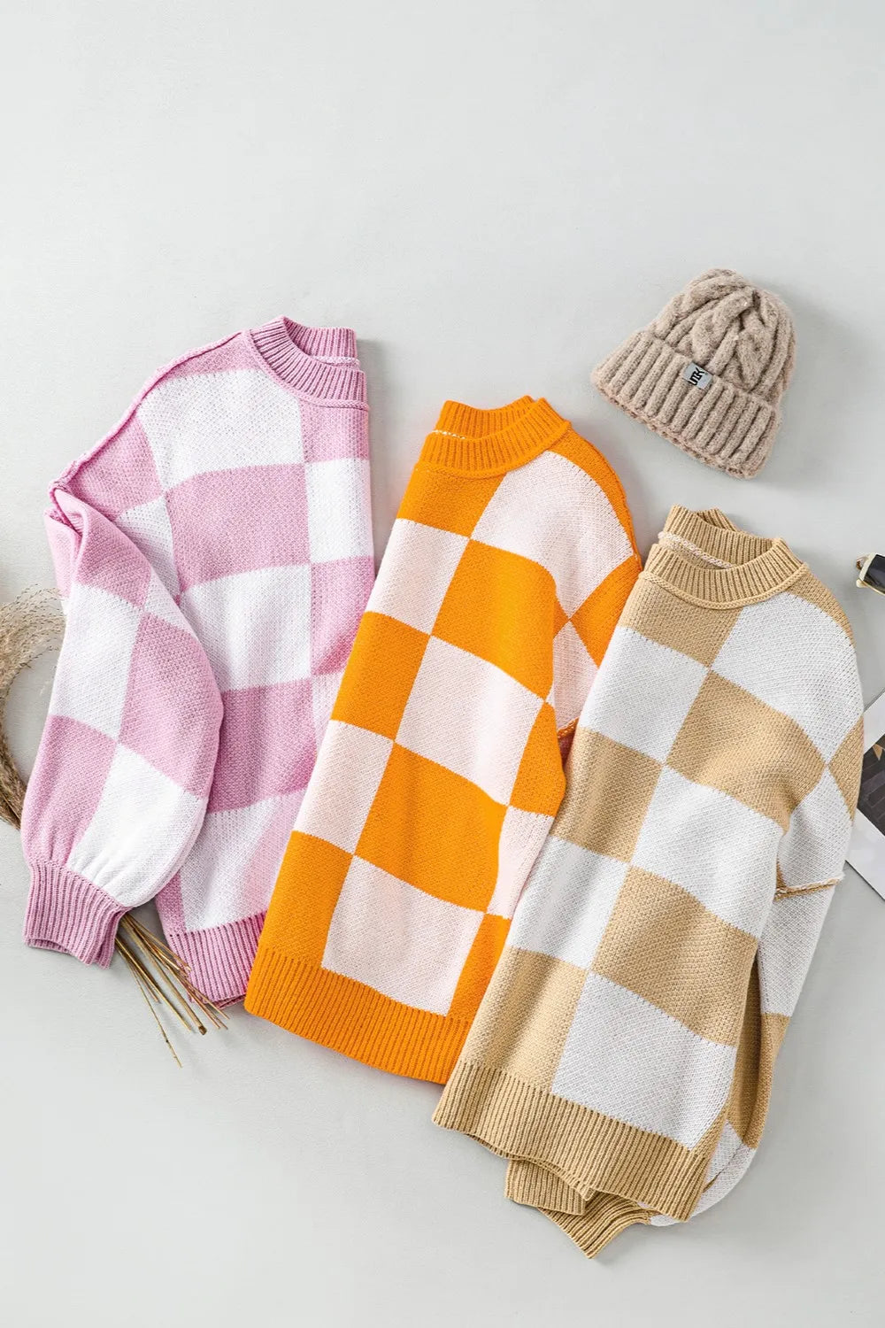 Checkered knit sweaters in pink, orange, and beige on hangers, crew neck and sleeves.