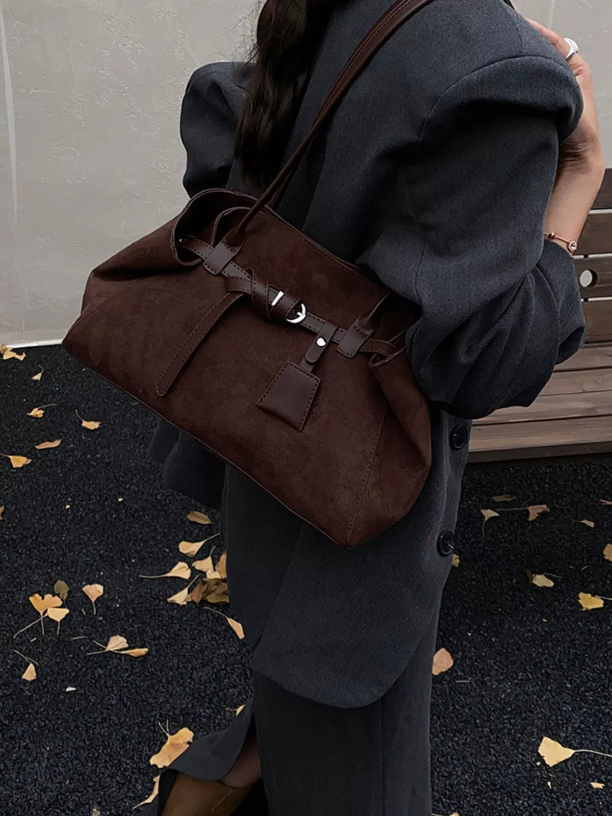 Brown suede shoulder bag with buckle strap and top handles, carried over dark coat