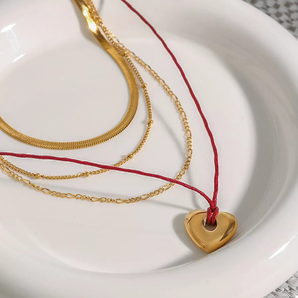 Gold layered heart necklace with red cord and heart pendant on white plate, close-up.