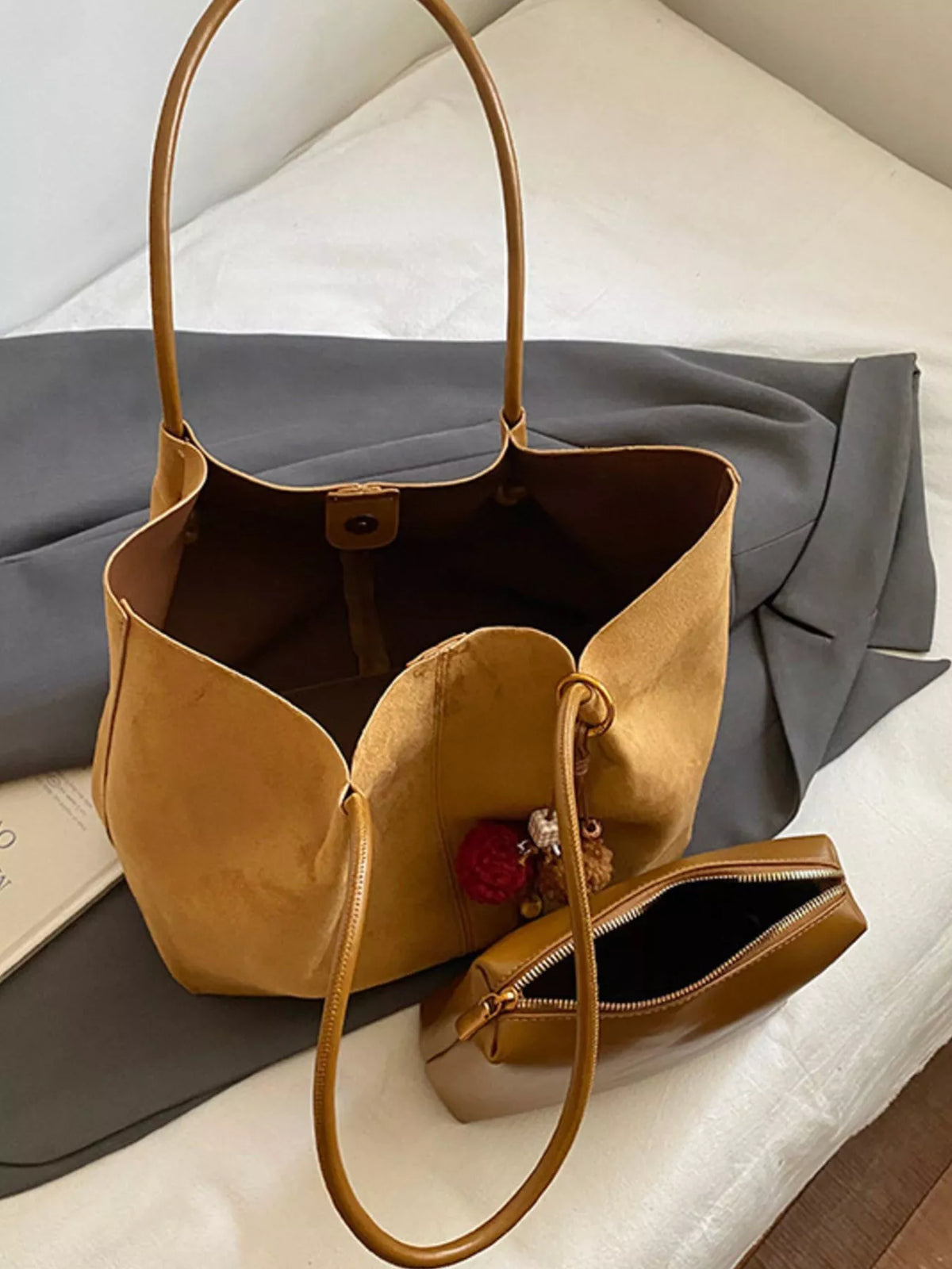 Tan suede open-top tote bag with long handles and interior lining visible; pom-pom charm.