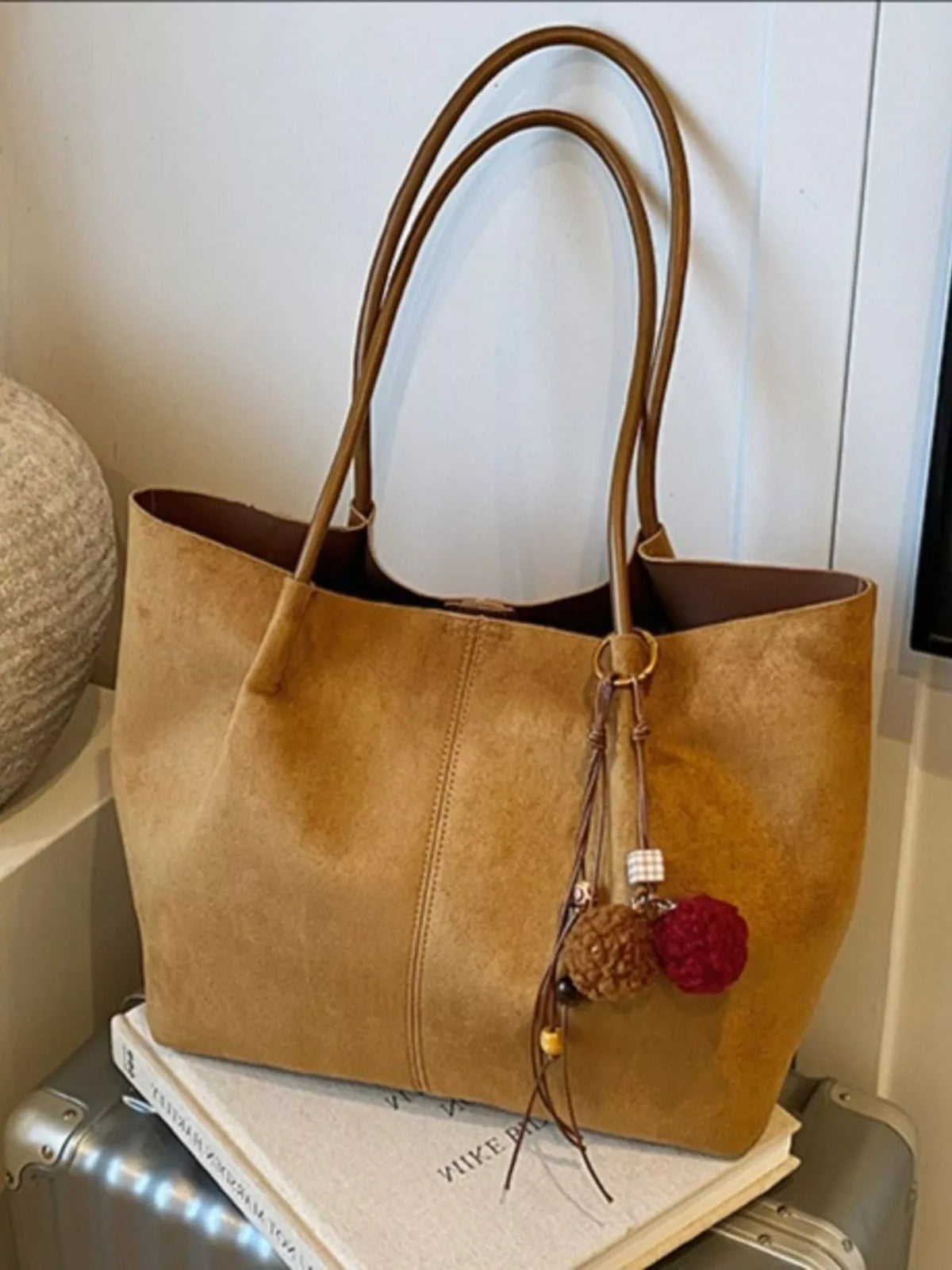 Tan suede tote bag with long handles, front zipper pocket, and pom-pom charm.