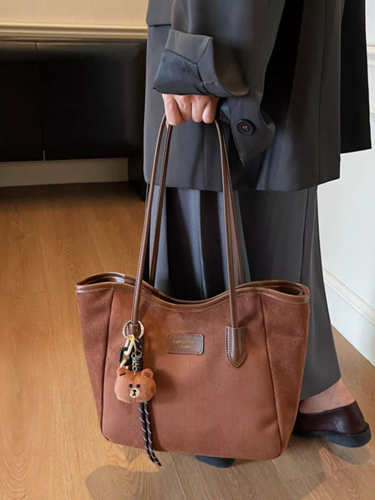 Brown suede tote bag with long straps, front logo patch, and bear keychain tassel