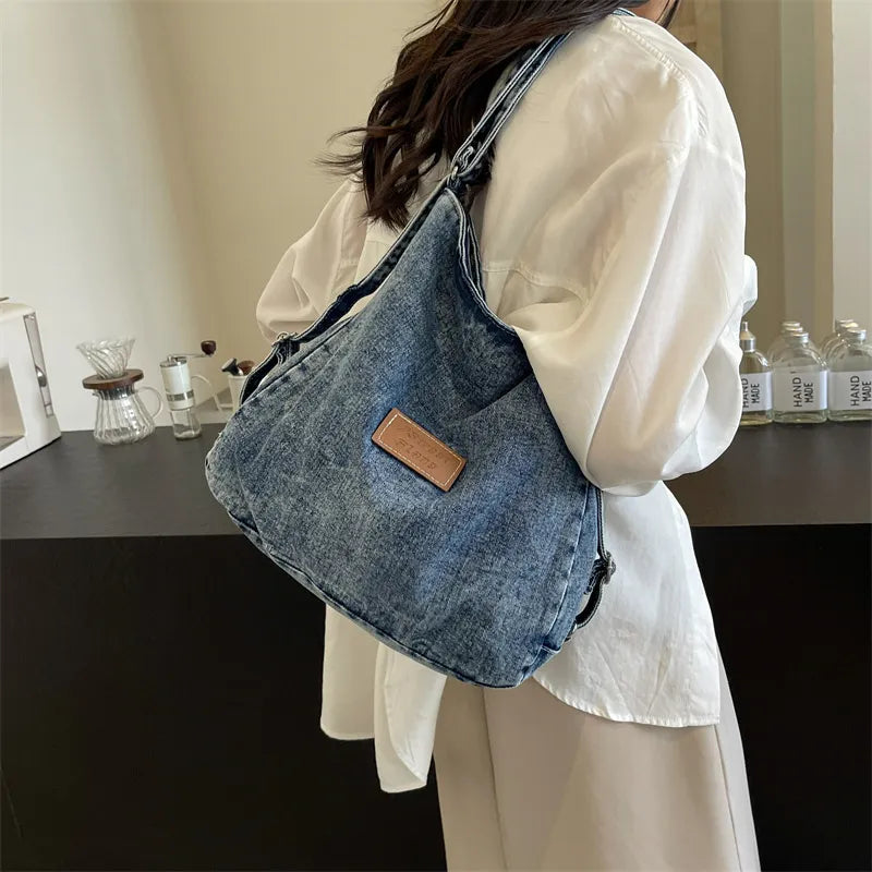 Adjustable strap denim shoulder bag worn on a person, blue fabric with brown logo patch