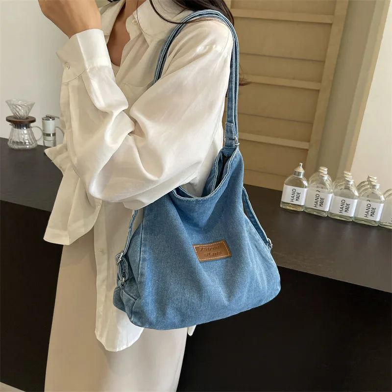 Adjustable strap denim shoulder bag worn on a person, medium blue with front brown patch