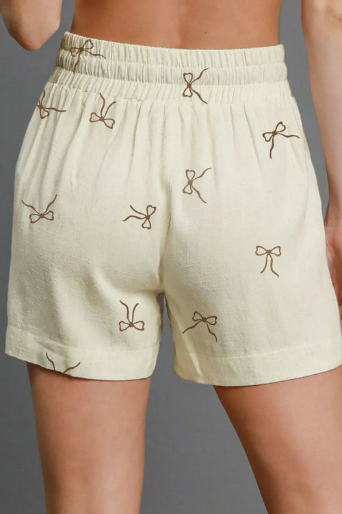 Cream shorts with elastic waistband, bow drawstring, and scattered brown bow prints