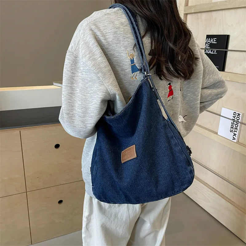 Dark blue denim shoulder bag worn crossbody with adjustable strap and logo patch